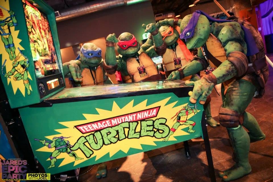 Costumed Ninja Turtles posing behind a Teenage Mutant Ninja Turtles pinball machine at the Epic Party