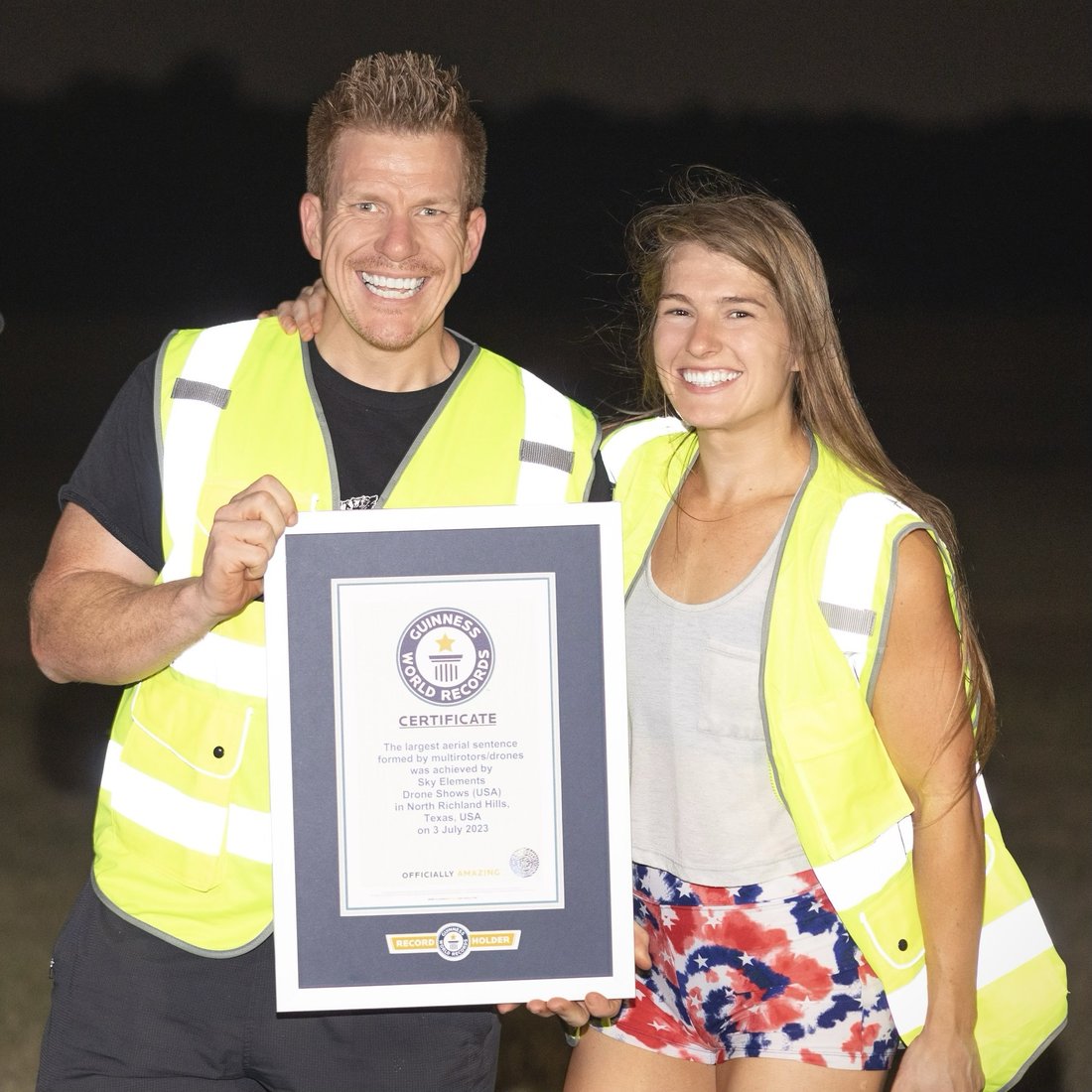 Jared Guynes and a colleague in hi-vis vests holding the certificate at the drone site at night