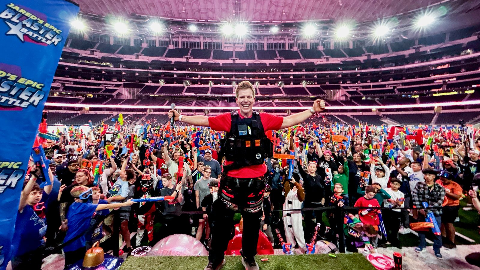 Jared Guynes on stage with his arms out at AT&T Stadium, with thousands of players raising their foam blasters behind him