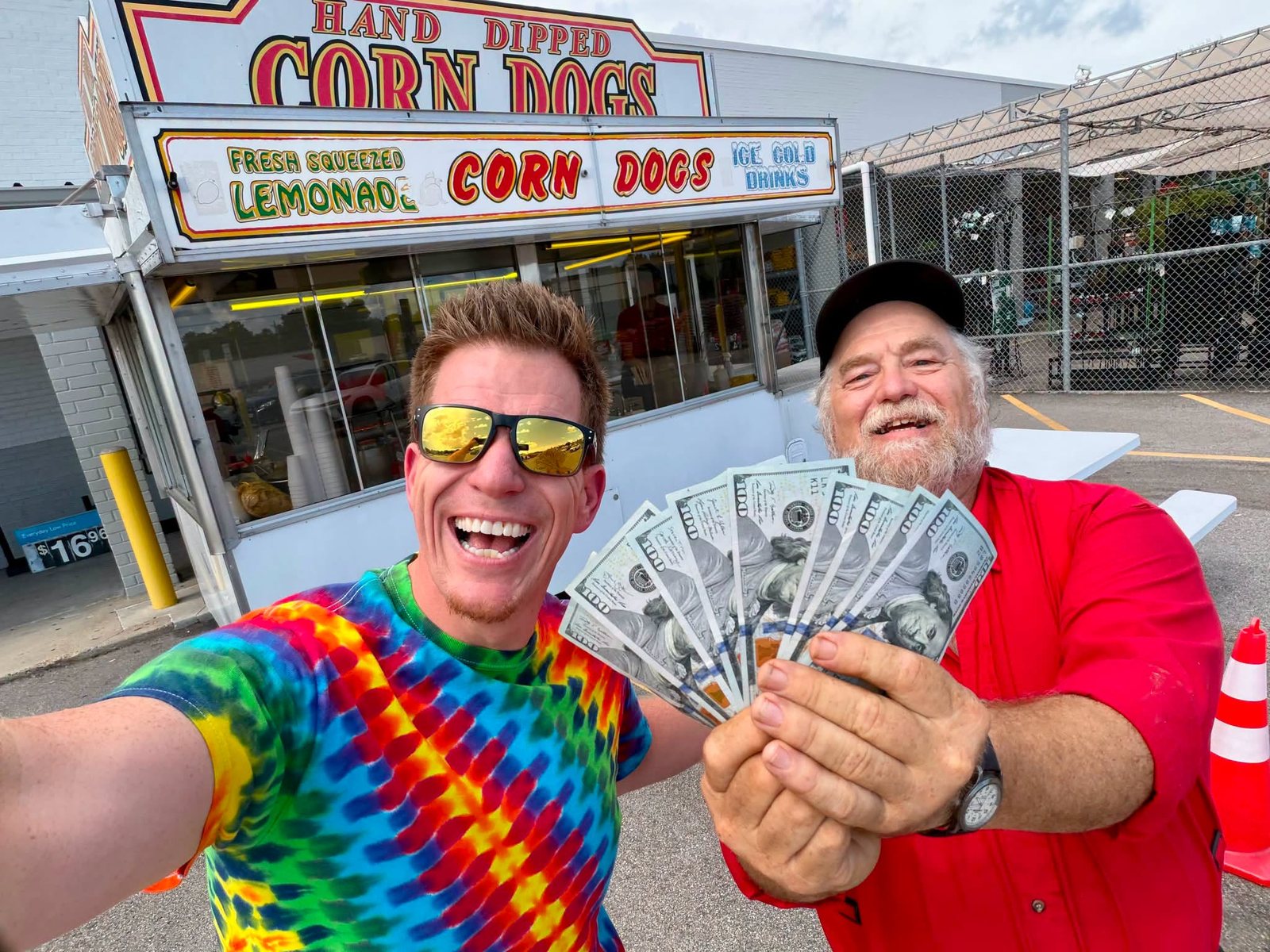 Jared Guynes holding a fan of hundred-dollar bills beside a man in a red shirt outside a hand-dipped corn dog stand